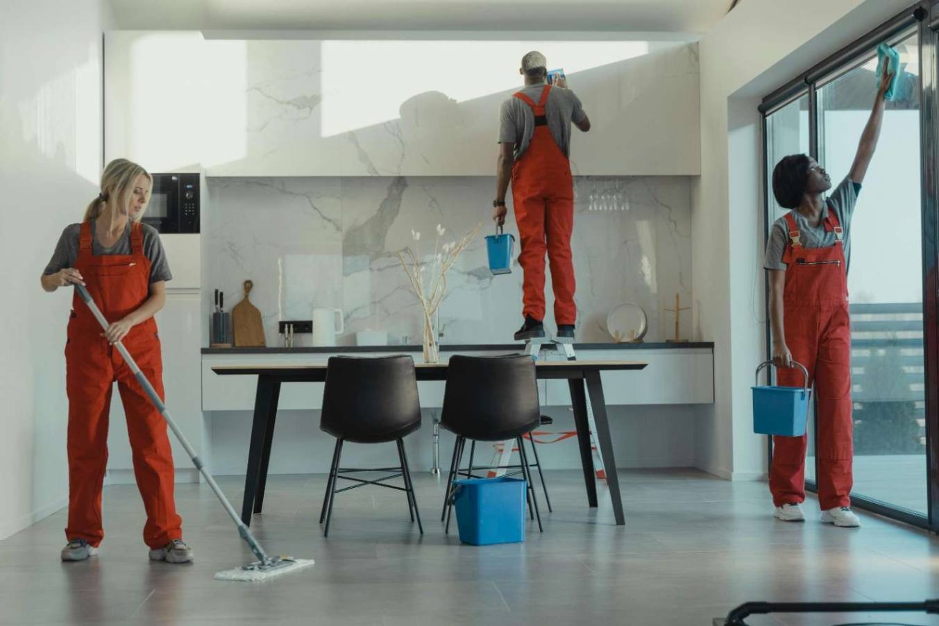 A team of three professional cleaners in bright orange overalls working in a modern, minimalist kitchen with marble accents. One person mops the floor, another cleans a high cabinet while standing on a step stool, and the third wipes a large glass sliding